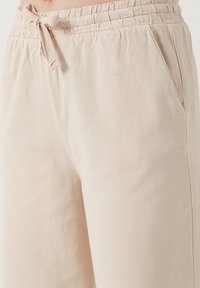 Light beige, textured pants with an elastic waistband, drawstring closure, and side pockets. Smooth fabric with subtle horizontal lines.