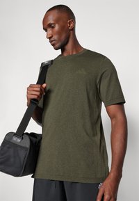 Camiseta corta de manga corta en verde mezclado con un escote redondo, vista en una persona que sostiene una bolsa negra con paneles de malla y sólidos.