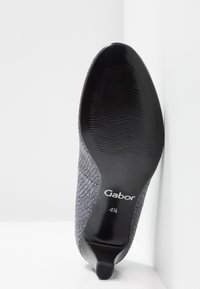 Chaussure à talon pour femme avec dessus texturé gris, semelle noire, et taille "Gabor 4½" imprimée en dessous, placée en angle contre un coin blanc.