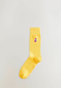 Chaussette jaune avec un petit logo brodé de canette de soda rouge et bleu sur la zone du mollet, posée à plat sur un fond clair.