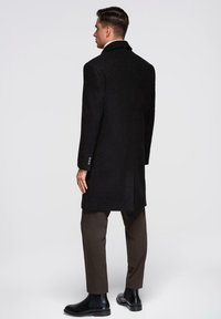 Schwarzer Wollmantel mit Reverskragen, tailliertem Schnitt und Seitentaschen, kombiniert mit braunen Hosen und schwarzen Stiefeletten.