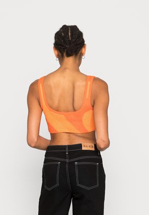 Femme vue de dos portant un crop top orange et un jean noir avec des coutures blanches et une étiquette "NA-KD" sur la ceinture.