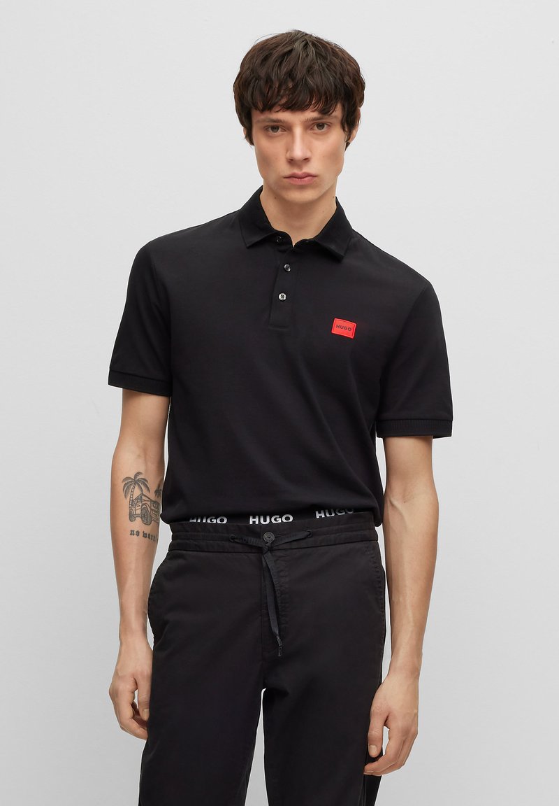 Black polo shirt with a red rectangular brand tag on the chest, paired with black drawstring trousers featuring a logo waistband.