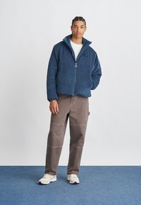 Giacca blu in corduroy con zip da uomo, t-shirt bianca sotto, abbinata a pantaloni marroni. Presenta un colletto alto e una toppa con logo.
