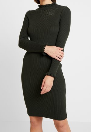 Robe côtelée vert foncé avec un col haut à volants. Manches longues avec des poignets à volants. Design ajusté, silhouette longueur genou.