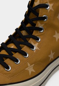 Kõrge säärega pruunast lõuendist tennisejalanõu mustade paelte, metallist silmuste, beežide tähemustrite ja kummist tallaga, millel on must triip.