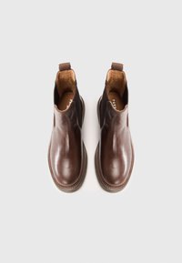 Des bottes Chelsea en cuir marron présentent des panneaux latéraux élastiques et une languette, avec un bout arrondi et des coutures le long de la semelle pour un détail supplémentaire.