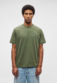 Camiseta de algodão verde-oliva com gola redonda, mangas curtas e um logotipo subtil em tinta mais escura no peito. Corte casual e textura suave.