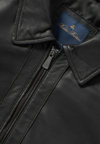 Černá kožená bunda s vysokým límcem, kovovým zipem a hladkým povrchem. Vnitřní štítek v námořnické modři se zlatým písmem.