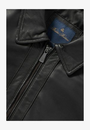 Schwarze Lederjacke mit hohem Kragen, Metallreißverschluss und glatter Oberfläche. Innenetikett in Marineblau mit goldenen Buchstaben.