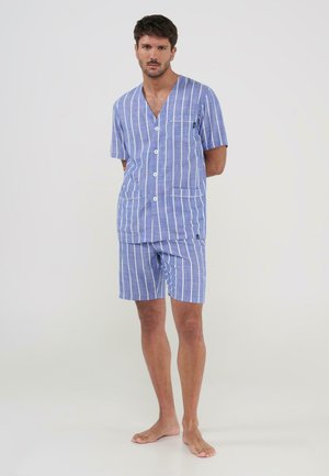 Hombre de pie descalzo vistiendo una camisa de manga corta con botones a rayas azules y blancas y pantalones cortos a juego sobre un fondo liso.