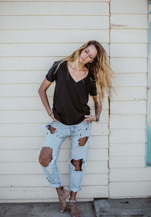 Kobieta z długimi blond włosami stoi boso przy jasnej drewnianej ścianie, ubrana w czarną koszulkę i mocno podarte, jasnoniebieskie jeansy, z rękami w kieszeniach.