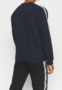 El sudadera de cuello redondo de marina presenta una franja lateral blanca con el logo, una textura suave y un corte relajado, combinada con pantalones de chándal negros que muestran acentos del logo.