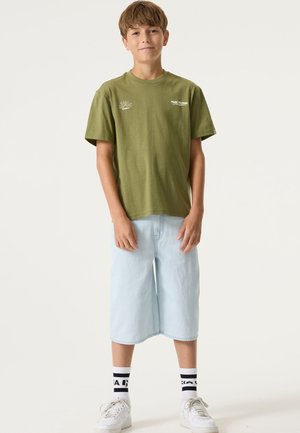 Maglietta a maniche corte verde oliva con logo, abbinata a pantaloncini azzurri fino al ginocchio e sneakers bianche, con calzini a righe nere e bianche.