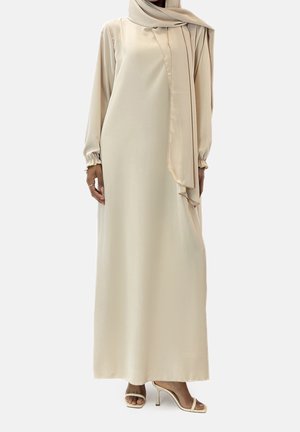 Kobieta w długiej beżowej sukience z długimi rękawami i dopasowanym hijabem, uzupełniona beżowymi sandałami na wysokim obcasie z odkrytymi palcami.