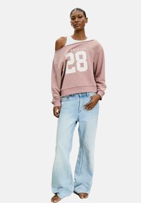 Felpa corta rosa con grafica bianca "28", design off-the-shoulder, abbinata a jeans a gamba larga azzurri, indossata a piedi nudi.