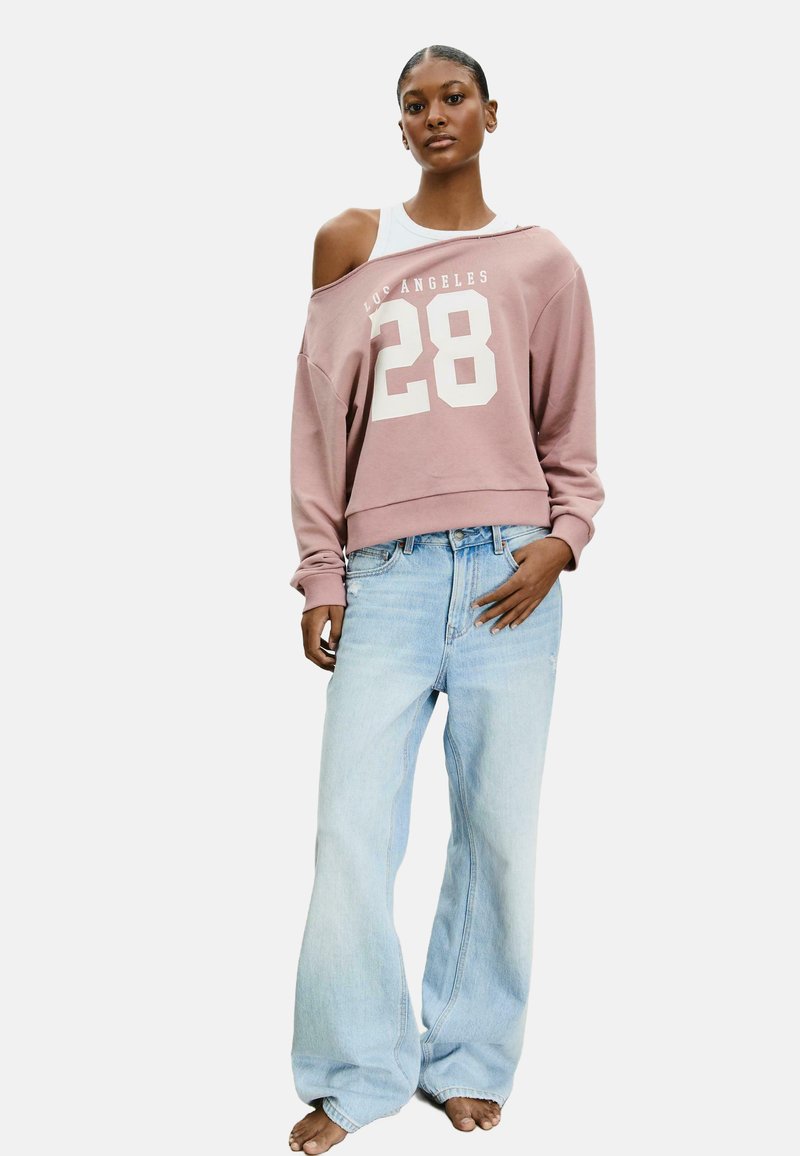 Felpa corta rosa con grafica bianca "28", design off-the-shoulder, abbinata a jeans a gamba larga azzurri, indossata a piedi nudi.