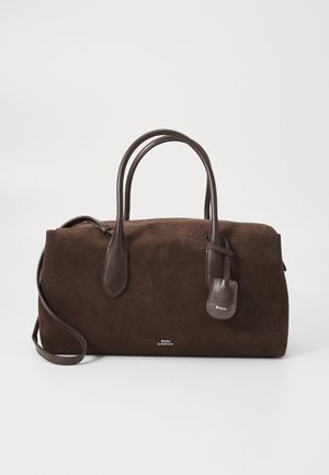 Sac en suède marron avec des poignées en cuir et une étiquette détachable, présenté sur fond blanc. Présente un design minimaliste.