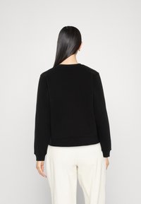 Donna con lunghi capelli neri che indossa un semplice felpa nera e pantaloni bianchi, in piedi di fronte a uno sfondo grigio chiaro.