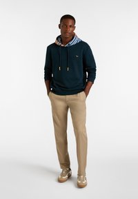 Felpa navy con fodera interna a righe multicolori, pantaloni beige e sneakers marrone chiaro. Presenta cordini e dettagli del logo frontale.