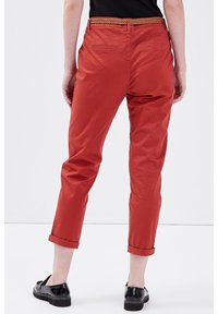 Pantalon en coton rouge avec une coupe effilée, revers retroussés et ceinture tissée. Deux poches arrière sont visibles. Une paire de chaussures noires complète la tenue.