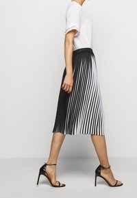 Femme portant un haut blanc à manches courtes, une jupe mi-longue plissée noir et blanc, et des sandales à talons noirs marchant de côté sur un sol gris.