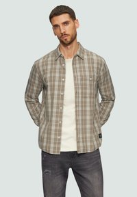 Chemise à carreaux marron et beige avec un col boutonné, des manches longues et une poche avant, portée par-dessus un T-shirt de couleur claire.