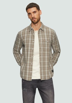 Chemise à carreaux marron et beige avec un col boutonné, des manches longues et une poche avant, portée par-dessus un T-shirt de couleur claire.