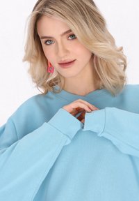 Svijetloplavi oversized dukserica s rebrastim rukavima i izrezom, s mekim teksturom. Model prikazuje opušteni kroj i vibrantne naušnice.