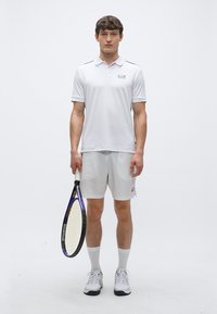 Balti polo marškinėliai su apykakle ir logotipu, derinami su šviesiai pilkomis šortais. Modelis laiko teniso raketę ir dėvi baltus sportinius batelius.