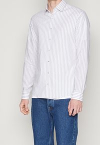 Bílá košile na knoflíky s tenkými vertikálními šedými pruhy, dlouhé rukávy s kontrastními pruhovanými manžetami a klasickým límcem. Kombinováno s modrými džínami.