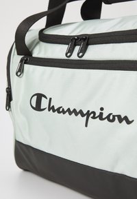 Svijetlo zeleni putni kofer s crnim detaljima, s dva crna patentna zatvarača i istaknutim crnim "Champion" logom.