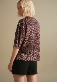 Tijelo smeđe majice s leopard uzorkom kratkih rukava, izrađeno od laganog materijala, u kombinaciji s crnim šortsevima. Vidljivi detalji šivanja.