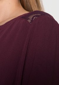 Haut bordeaux présentant un tissu texturé avec des accents en dentelle à l'épaule, affichant une coupe douce et drapée et un col arrondi.