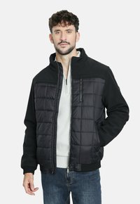 Hombre vestido con una chaqueta acolchada negra sobre un suéter blanco con cremallera y jeans azules, de pie con una mano en el bolsillo contra un fondo blanco.