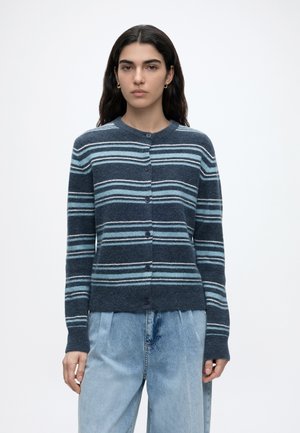 Femme aux longs cheveux foncés porte un cardigan bleu marine boutonné avec des rayures horizontales bleu clair et un jean bleu clair ample.
