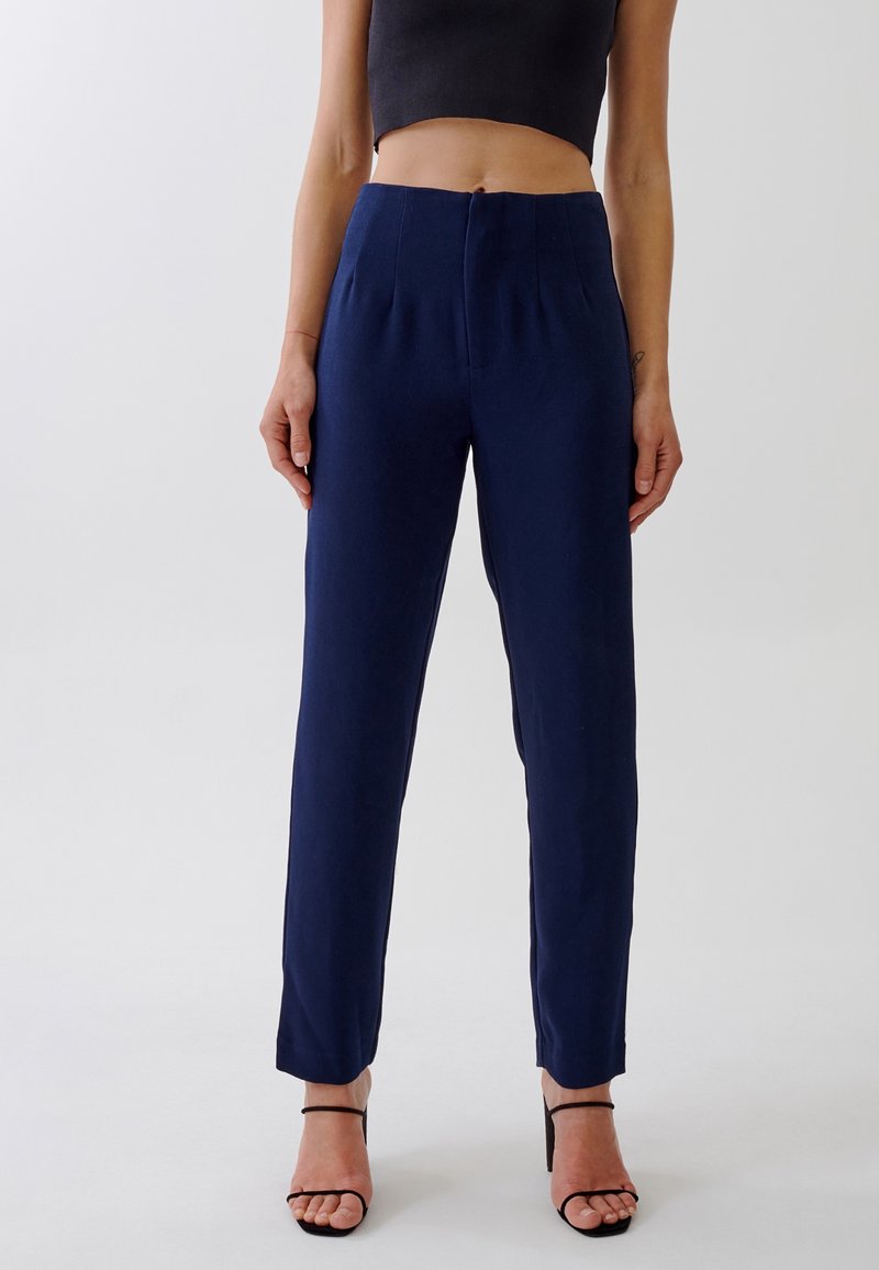 Femme portant un pantalon bleu marine taille haute ajusté, un top court noir et des sandales noires à talons ouverts, debout devant un fond uni.