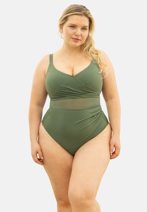 Frau mit langen blonden Haaren, die einen olivgrünen Badeanzug mit Netzbesatz und geraffter Taille trägt und vor einem einfachen Hintergrund steht.