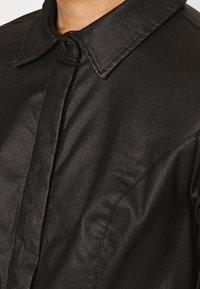 Czarna kurtka koszulowa o błyszczącej powierzchni, z ostrym kołnierzykiem, subtelnymi detalami zszycia i zapięciem na guziki z przodu.