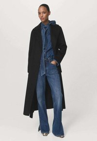 Long manteau noir sur une tenue en denim bleu, comprenant une veste à capuche et un jean évasé. Le mannequin se tient avec les mains dans les poches.