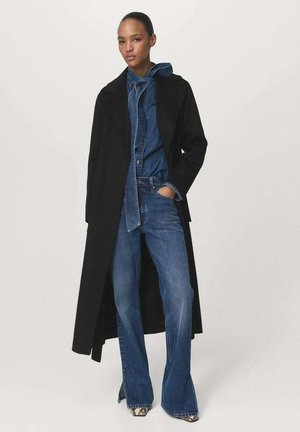 Lungo cappotto nero su un outfit in denim blu, caratterizzato da una giacca con cappuccio e jeans svasati. Il modello sta in piedi con le mani nelle tasche.