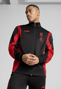 Must musta ja punane sportlik jakk, millel on lukuga sulgemine, Puma logo ja jalgpalli bränding. Kanga pind näeb välja siledana, elastsed mansetid ja allserv.