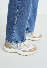 Hvite og beige joggesko med mesh- og skinnakenter, med en kraftig såle og reflekterende detaljer, kombinert med blå denim jeans.