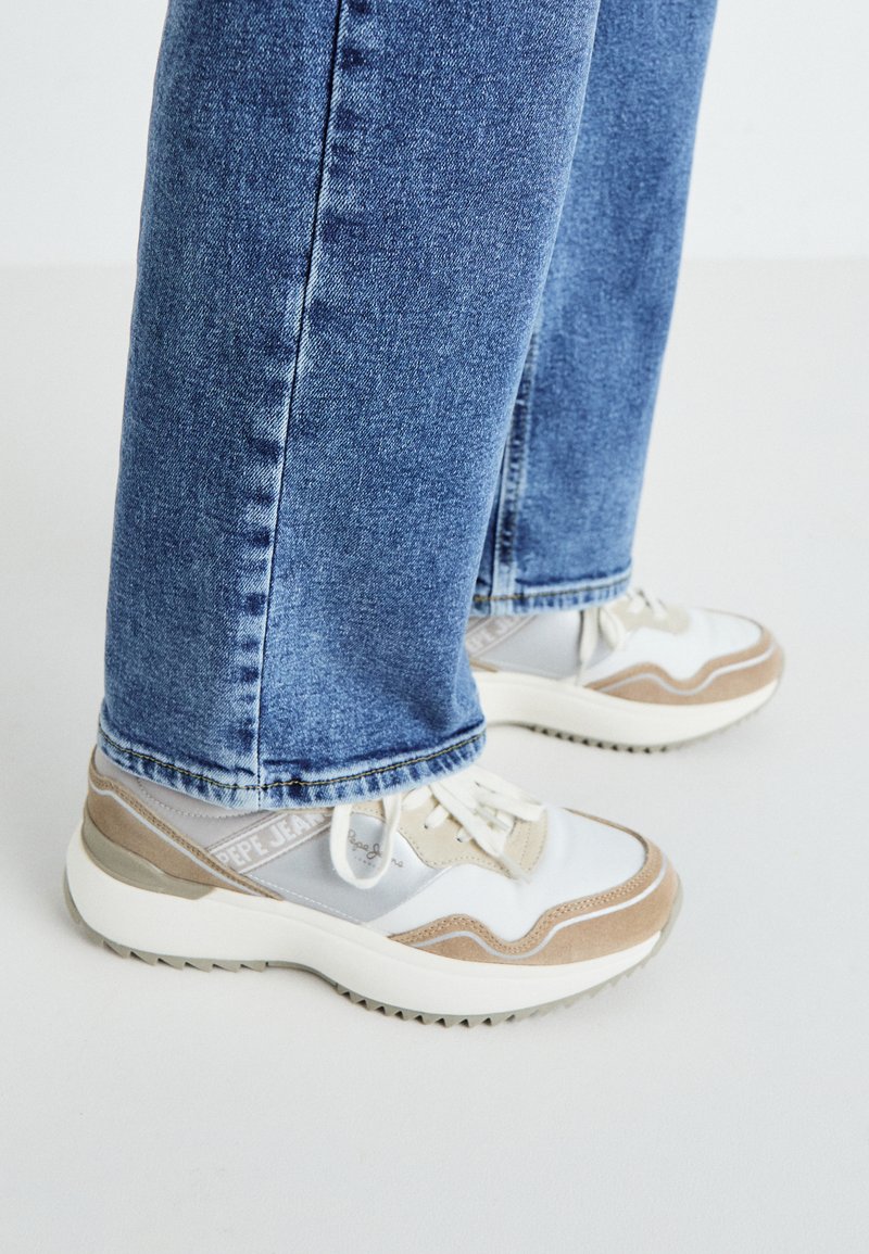 Zapatillas deportivas blancas y beige con acentos de malla y ante, que presentan una suela gruesa y detalles reflectantes, combinadas con jeans de mezclilla azul.