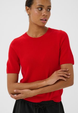 Femme portant un pull rouge vif à manches courtes, les bras croisés, regardant sur le côté sur un fond clair uni.