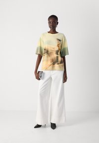 Loose-fitting, short-sleeved t-shirt in soft fabric, featuring an abstract green and yellow print. Paired with wide white trousers and black heels.