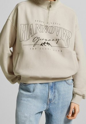 Beżowa bluza z ćwierćzamkiem z napisem "Hannover Niemcy" i grafiką gór, noszona z jasnoniebieskimi dżinsami, z ręką swobodnie opartą wzdłuż ciała.