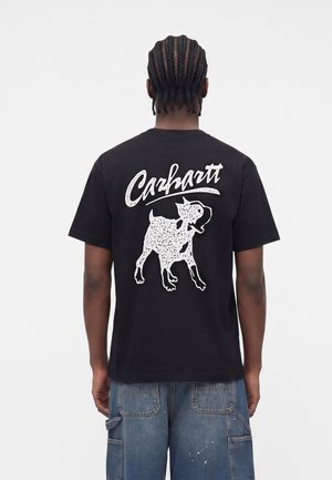 Mężczyzna z zaplecionymi włosami, ubrany w czarną koszulkę Carhartt z białą grafiką psa i logo na plecach, w połączeniu z niebieskimi dżinsami.