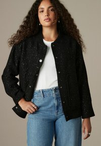 Svart texturerad jacka med silverdetaljer, rund halsringning, knappstängning och framficka, kombinerad med en vit skjorta och blå jeans.