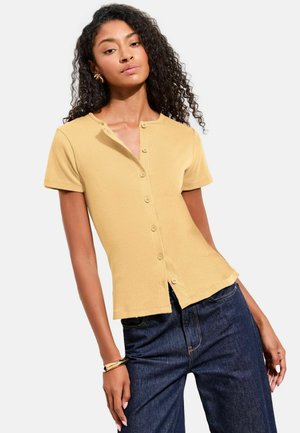 Frau mit lockigem Haar, die ein gelbes kurzärmeliges Hemd mit Knöpfen und dunkelblaue Jeans trägt und mit einer Hand in der Hüfte posiert.